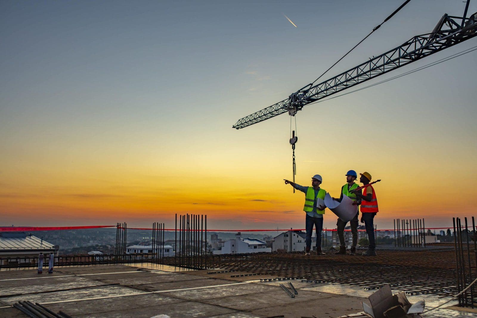 Engineers in hard hats reviewing plans beneath a tower crane on a construction site at sunset, as JLL raises its Saudi tender price inflation forecast
