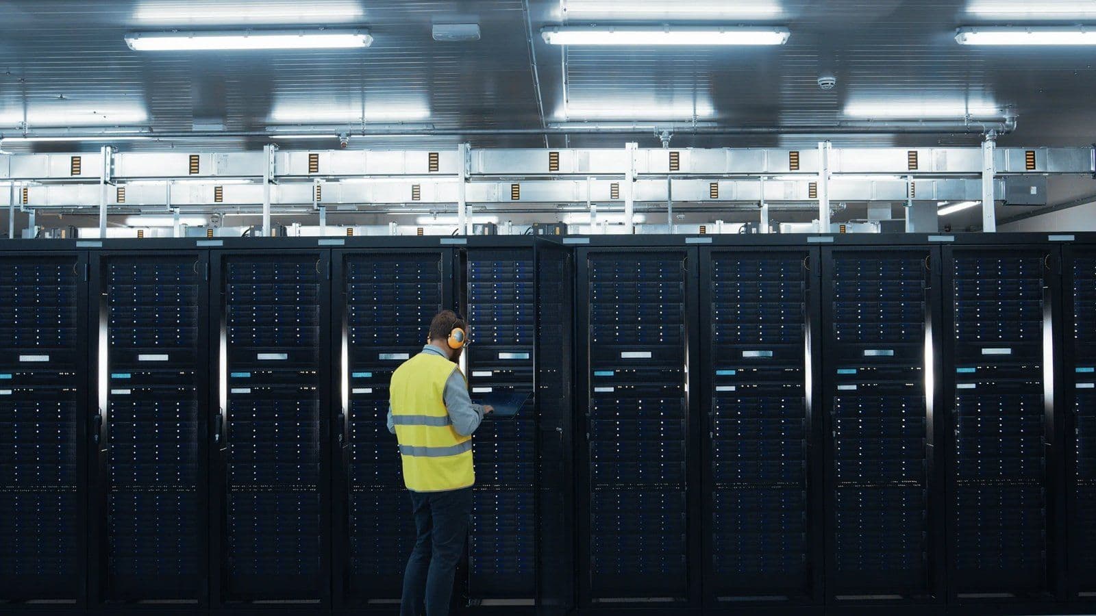 Engineer in a high-visibility vest working on a laptop beside rows of server racks in a data centre